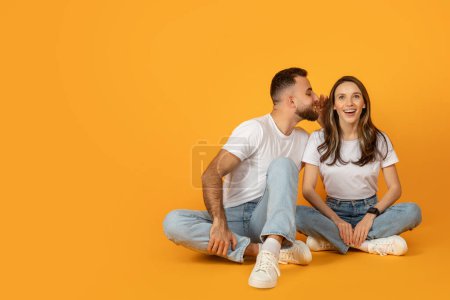 Téléchargez les photos : Un jeune couple joyeux en tenue décontractée s'assoit les jambes croisées sur un fond orange vif, avec l'homme chuchotant amusamment dans l'oreille des femmes, la faisant rire - en image libre de droit
