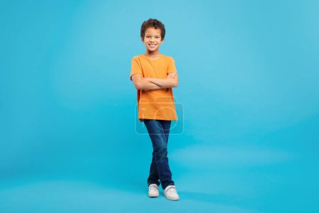 Téléchargez les photos : Jeune écolier confiant avec un sourire amical, les bras croisés, portant un t-shirt orange et un jean sur fond bleu apaisant, pleine longueur - en image libre de droit