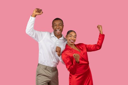 Foto de Hombre negro exuberante en camisa blanca y mujer extática en vestido rojo levantando los puños en celebración, irradiando felicidad y victoria sobre fondo rosa - Imagen libre de derechos