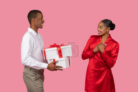 Foto de Hombre negro generoso en camisa blanca presentando gran regalo con arco a la mujer tocada en vestido rojo, sus manos en su corazón, sobre fondo rosa tierno - Imagen libre de derechos