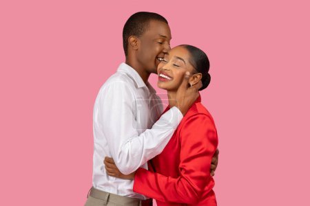 Photo for Joyful black man in white shirt affectionately kissing cheek of smiling woman in red dress, sharing tender moment against pink background - Royalty Free Image