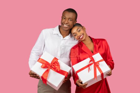 Téléchargez les photos : Couple noir radieux tenant chacun une boîte cadeau blanche attachée avec un ruban rouge, homme vêtu d'une chemise blanche et femme en robe rouge, les deux souriant de joie sur fond rose - en image libre de droit