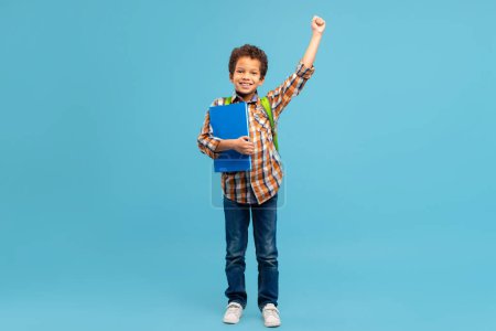 Photo for Victorious young boy in checkered shirt with raised fist, holding blue book and wearing backpack, stands against bright blue background, full length - Royalty Free Image