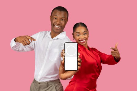 Photo for Excited black couple presenting smartphone pointing at blank screen, showcasing place for your design, app or ad with enthusiasm on pink backdrop, mockup - Royalty Free Image