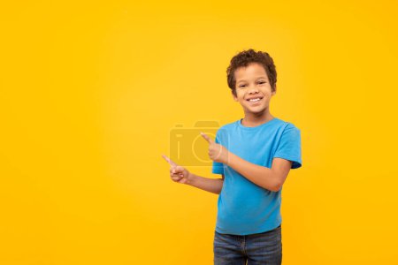 Téléchargez les photos : Joyeux garçon latin aux cheveux bouclés pointant vers son côté avec un grand sourire, montrant l'espace de copie, portant un t-shirt bleu, sur fond jaune uni - en image libre de droit