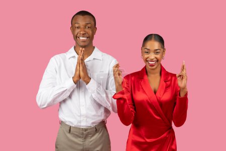 Foto de Hombre negro optimista en camisa blanca con las manos en pose de oración y mujer alegre en vestido rojo cruzando sus dedos, ambos sonriendo con esperanza y positividad contra el telón de fondo rosa - Imagen libre de derechos