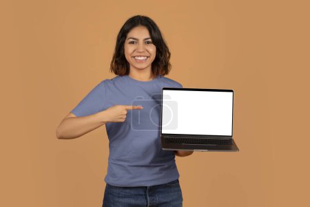 Téléchargez les photos : Joyeux jolie jeune femme arabe montrant ordinateur portable avec écran blanc vierge, recommandant beau site Web éducatif, offre en ligne pour les étudiants, espace de copie, maquette, fond brun - en image libre de droit
