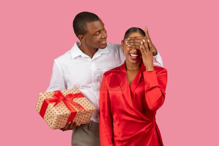 Foto de Hombre negro con camisa blanca sostiene el regalo detrás de su espalda mientras cubre juguetonamente los ojos de la mujer usando un vestido rojo, preparando la sorpresa sobre el fondo rosa - Imagen libre de derechos
