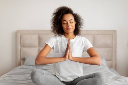 Téléchargez les photos : Calme jeune femme noire méditant en position lotus avec les mains jointes ensemble, relaxant pendant le yoga du matin, assis sur le lit à l'intérieur de la chambre à coucher à la maison, embrassant pleine conscience et détente - en image libre de droit