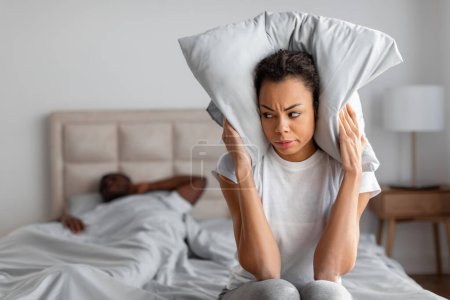 Photo for Unhappy young african american lady covering ears with pillow on bed, suffering from husband snoring, sitting unable to sleep in modern bedroom indoor. Noisy neighbors concept - Royalty Free Image