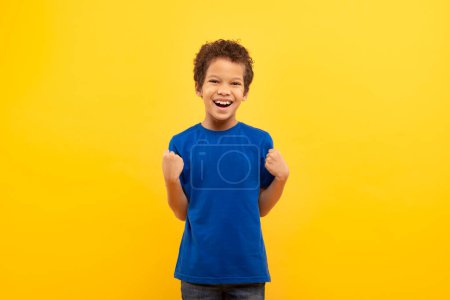 Téléchargez les photos : Garçon exubérant aux cheveux bouclés riant et serrant les poings dans la pose de la victoire, célébrant le succès, portant un t-shirt bleu royal sur fond jaune - en image libre de droit