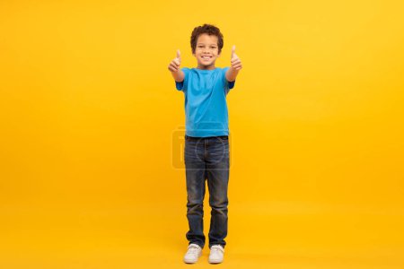 Foto de Niño feliz con el pelo rizado dando pulgares dobles hacia arriba, vistiendo camiseta azul, lleno de entusiasmo, contra el fondo amarillo soleado, longitud completa - Imagen libre de derechos