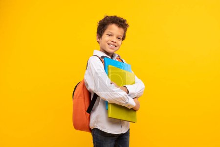Téléchargez les photos : Écolier confiant aux cheveux bouclés, tenant des livres colorés et portant un sac à dos, prêt pour l'école sur fond jaune vif - en image libre de droit