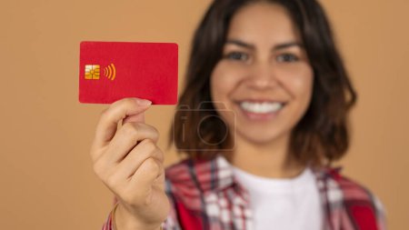 Photo for Selective focus on red plastic credit card in smiling young arab woman hand, isolated on brown studio background. Easy banking, contactless payment, virtual shopping concept - Royalty Free Image