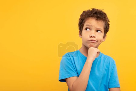 Téléchargez les photos : Écolier pensif aux cheveux bouclés tenant son menton, portant un t-shirt bleu vif, avec une expression quiz regardant l'espace libre sur fond jaune - en image libre de droit