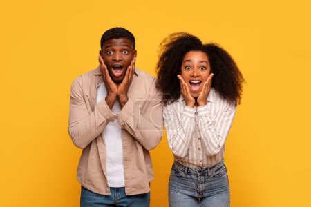 Photo for Surprised black man and woman with hands on their cheeks, wide-eyed and open-mouthed in astonishment, standing together against vibrant yellow background - Royalty Free Image