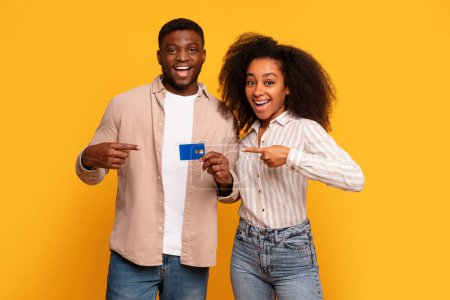 Foto de Sonriente joven pareja negra señalando con entusiasmo a la tarjeta de crédito que tienen, sugiriendo aprobación financiera o nueva compra, contra el soleado fondo amarillo - Imagen libre de derechos
