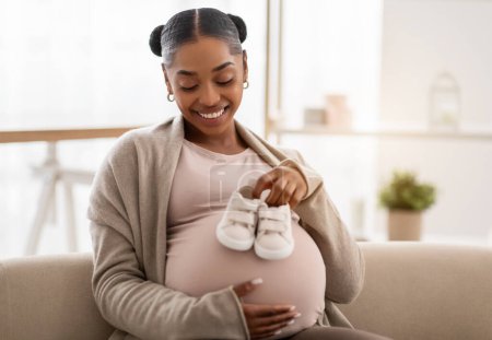 Photo for Waiting for a child, pregnancy. Joyful beautiful pregnant young african american woman sitting on couch at home, holding cute little baby booties on her big tummy and smiling, copy space - Royalty Free Image