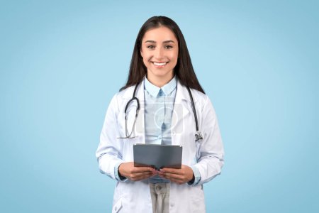 Téléchargez les photos : Médecine européenne en manteau blanc, tenant une tablette numérique et souriant à la caméra, symbolisant les soins de santé modernes, posant sur fond bleu - en image libre de droit