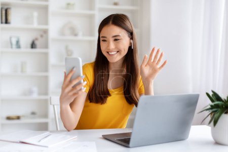 Téléchargez les photos : Joyeux jeune asiatique femme faire appel vidéo sur Smartphone tout en étant assis au bureau dans le bureau à domicile, heureuse excitée coréenne profiter de la téléconférence, ayant pause dans le travail, gros plan - en image libre de droit