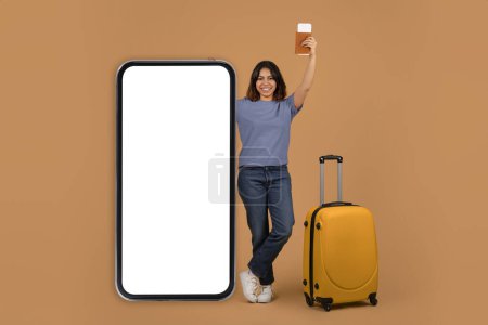 Photo for Pretty arab young woman tourist holding passport with travel tickets, stand next to big smartphone with mockup empty screen, standing on colorful background, great travelling offer, online checkin - Royalty Free Image