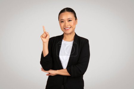 Photo for Confident Asian businesswoman smiling and pointing upwards with one hand, while the other arm is folded, showing a gesture of idea or solution in a professional environment - Royalty Free Image