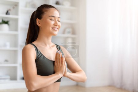 Foto de Hermosa mujer asiática practicando yoga con los ojos cerrados y haciendo gesto namaste, sonriendo tranquila joven coreana expresando paz y gratitud, haciendo yoga en su hogar minimalista soleado - Imagen libre de derechos