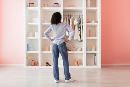 Photo for From behind, woman with curly hair stands with her hands on her hips, contemplating her well-organized wardrobe in a room with pink walls - Royalty Free Image