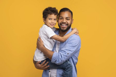 Photo for Happy black father lifting his little son and hugging, happy african american family of three smiling joyfully at camera, posing together against sunny yellow background, copy space - Royalty Free Image
