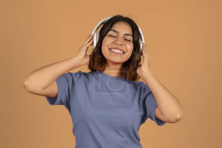 Photo for Cute positive funny young arab woman wearing casual clothing listening to music and dancing, raising hands up and smiling, using modern wireless headphones, enjoying favorite song, brown background - Royalty Free Image