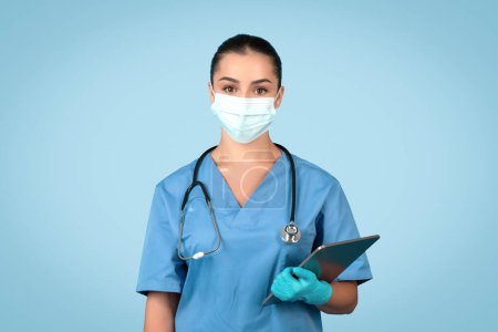 Foto de Doctora confiada, enfermera en uniforme azul, máscara protectora y guantes sujetando portapapeles, posando aislada sobre fondo azul del estudio. Medicina, atención médica, tratamiento - Imagen libre de derechos
