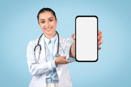 Téléchargez les photos : Médecin femelle européenne montrant un énorme smartphone avec écran blanc et sourire, médecin démontrant téléphone vide avec espace de copie pour la publicité, maquette - en image libre de droit