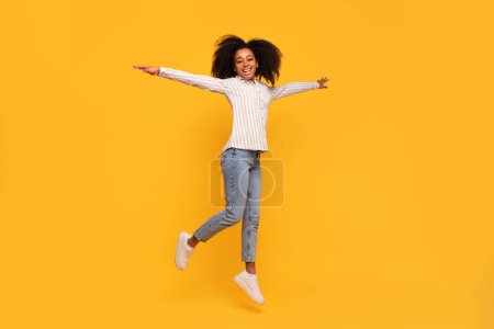 Photo for Radiant young black woman with arms wide open joyfully leaps against mustard yellow background, sporting casual chic with striped shirt and jeans - Royalty Free Image