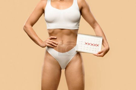 Photo for Unrecognizable young woman in white underwear holding calendar with marked days, having period or experiencing menstruation delay, standing isolated on beige studio background, free space - Royalty Free Image