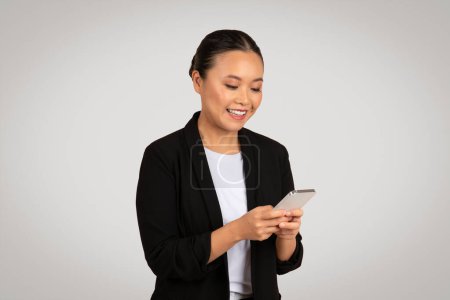 Foto de Mujer de negocios asiática sonriente con un traje negro absorto en su teléfono inteligente, posiblemente leyendo un buen mensaje de noticias o entablando una agradable conversación digital, aislado en un fondo gris, estudio - Imagen libre de derechos