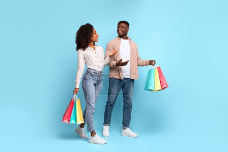 Photo for Relaxed, casually dressed black couple enjoys light conversation while holding vibrant shopping bags, set against soothing blue backdrop, evoking sense of leisure and contentment - Royalty Free Image