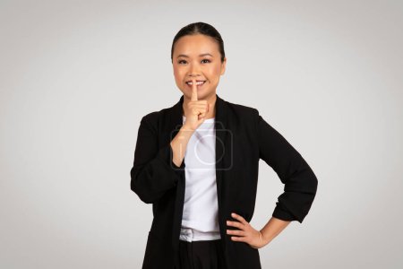 Photo for Professional Asian businesswoman with a secretive expression placing a finger to her lips in a hushing gesture, implying confidentiality or silence, posed against a grey background - Royalty Free Image