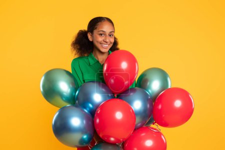 Téléchargez les photos : Joyeux adolescent noir fille posant avec un tas de ballons festifs colorés, exprimant la joie de fête d'anniversaire et de fête dans un studio jaune vibrant, souriant à la caméra - en image libre de droit