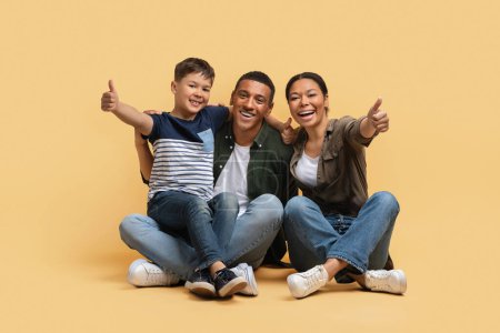 Photo for Funny cute family father mother son have fun isolated on beige studio background, sitting on floor, showing thumb ups and smiling at camera. Family offer, deal concept - Royalty Free Image