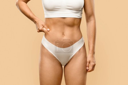 Photo for Slim young woman in underwear pinching fat of her waist zone, unrecognizable female with fit body ready for lypolisis or liposuction treatment, standing over beige studio background, copy space - Royalty Free Image
