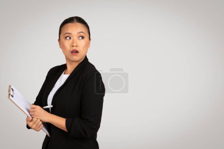 Foto de Empresaria asiática sorprendida en un traje negro con un portapapeles reacciona con una expresión de ojos abiertos, la captura de un momento de acontecimientos empresariales inesperados, aislado en el fondo gris, estudio - Imagen libre de derechos