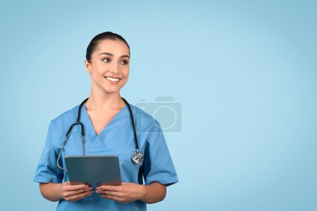 Cheerful young woman nurse in blue medical uniform holding digital tablet and looking thoughtfully aside at free space against blue background, place for medical ad