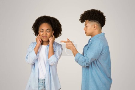 Eine frustrierte afrikanisch-amerikanische junge Frau hält sich die Ohren zu, um einen jungen Mann zu ignorieren, der auf sie zeigt und in einem neutralen Hintergrund ein übliches Argument oder ein Unstimmigkeitsszenario darstellt.