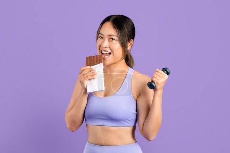 Téléchargez les photos : Souriant jeune femme asiatique en vêtements de sport tient la barre de chocolat dans une main et haltère dans l'autre, mélangeant indulgence avec fitness, fond violet - en image libre de droit