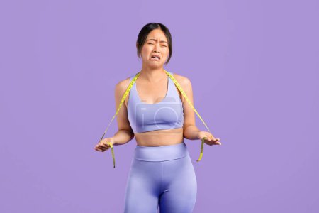 Upset Asian woman in sportswear wearing measuring tape around her neck and crying, expressing frustration with body image and fitness goal, purple backdrop
