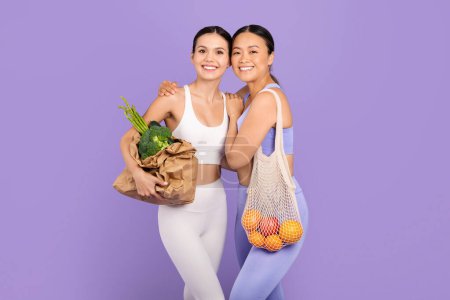 Téléchargez les photos : Deux femmes diverses en tenue d'entraînement, l'une avec sac de légumes biologiques, l'autre avec sac net de fruits, présentant des options alimentaires saines sur fond violet - en image libre de droit