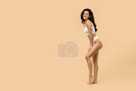 Photo for Smiling beautiful woman with sculpted figure posing in white lingerie against beige background, happy young female with fit shape embodying health and body care in neutral studio backdrop - Royalty Free Image