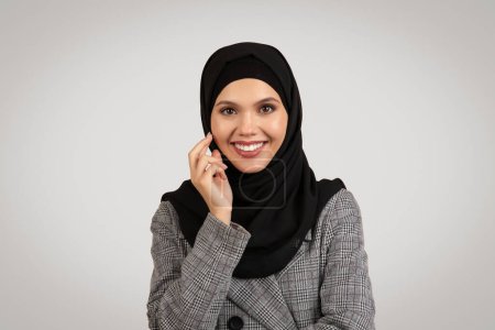 Téléchargez les photos : Portrait de femme d'affaires musulmane en hijab souriant à la caméra, posant élégamment sur fond de studio gris, symbolisant la grâce et la fierté culturelle - en image libre de droit