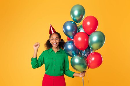 Téléchargez les photos : Heureuse adolescente afro-américaine en chapeau d'anniversaire festif posant avec un tas de ballons colorés, geste oui célébrer sa journée spéciale sur fond de studio jaune - en image libre de droit