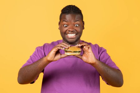 Photo for Cheat Meal. Portrait Of Hungry Black Guy In Purple T-Shirt Eating Yummy Burger, Holding And Looking At Tasty Junk Food, Enjoying Snack Over Orange Yellow Studio Background - Royalty Free Image